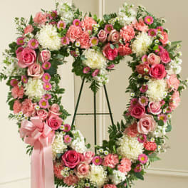 Heart-shaped standing wreath of pink roses, carnations, and white mums with a pink ribbon bow.
