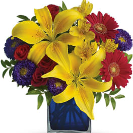 Bright mixed arrangement with yellow lilies, red roses and gerberas, and purple asters in a blue glass cube vase