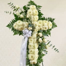 Standing cross-shaped spray of white flowers and roses with a white ribbon on an easel.