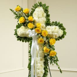 Tall floral cross of white flowers edged in green with yellow roses and a white ribbon on a standing easel