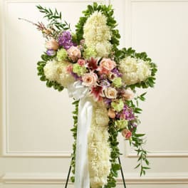 Tall standing white floral cross with pastel roses and lilies on an easel with white ribbon.