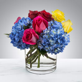 Low arrangement of red, pink, and yellow roses with blue hydrangeas in a clear glass vase