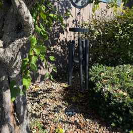 Black memorial wind chime with hummingbird design hanging from a tree over a rock garden.