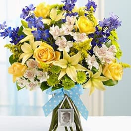 Mixed bouquet of yellow roses, lilies, and blue flowers in a glass vase with a baby photo charm.