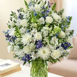 Tall vase arrangement of white roses and mums with blue accent flowers in a clear glass cylinder