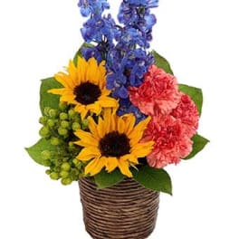 Bright arrangement with sunflowers, blue delphinium, and pink carnations in a small wicker basket