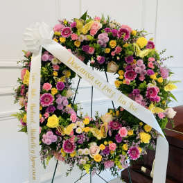 Large floral wreath with pink, yellow, and purple blooms on a stand with white ribbons