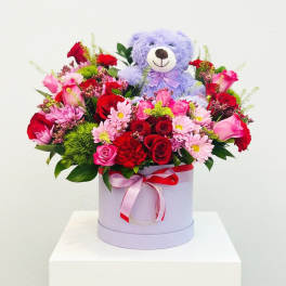 Lavender teddy bear in a box of pink and red flowers