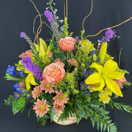Basket arrangement with peach roses, yellow lilies, purple spikes, and blue flowers.