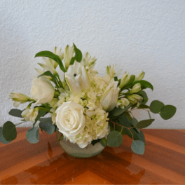 Low white arrangement of roses, lilies, and hydrangeas in a round glass vase