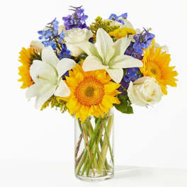 Bouquet of white lilies, sunflowers, roses, and blue flowers in a glass vase