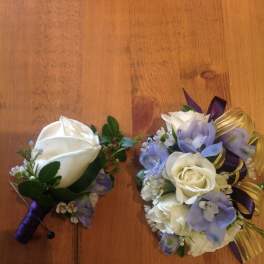 Two small floral corsages with white roses and purple accents on a wooden surface.