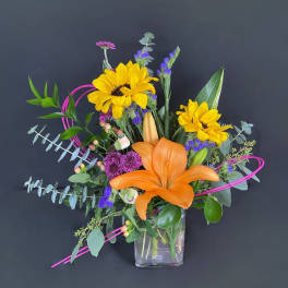 Bright mixed bouquet with sunflowers, orange lily, and purple accents in a glass vase