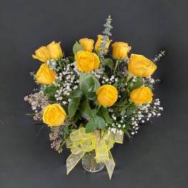 Arrangement of yellow roses with white filler flowers in a clear glass vase with a yellow bow.