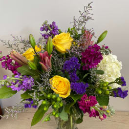 Mixed bouquet of yellow roses, purple flowers, and white hydrangea in a glass vase