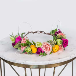 Colorful flower crown with pink roses, yellow blooms and green buds on a vine base, displayed on a round marble table.