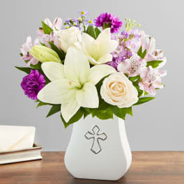Bouquet of white lilies, roses, and purple flowers in a white vase with a cross