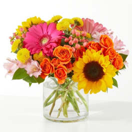 Mixed bouquet of gerbera daisies, roses, and sunflowers in a glass vase