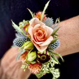 Wrist corsage with a peach rose, spiky blue blooms, and orange accents on a person’s wrist
