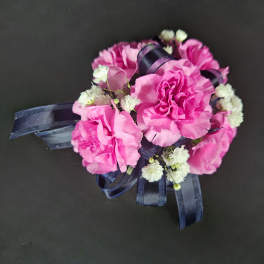 Pink carnation corsage with white filler flowers and navy ribbon on a dark background