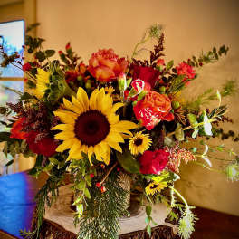 Rustic arrangement of sunflowers, red and orange roses, and daisies in a wood-textured vase