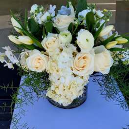 Low arrangement of ivory roses, tulips, orchids, and hydrangeas in a glass vase with trailing greenery