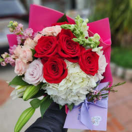 Handheld bouquet of red roses, pink blooms, white hydrangea and lilies with a lavender envelope and pink wrap