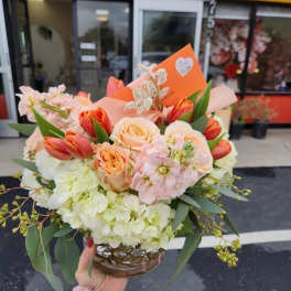 Low bouquet of peach roses, orange tulips, and white hydrangeas in a clear round vase with an orange card.