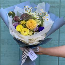 Hand-tied bouquet of yellow, purple, and white flowers wrapped in blue paper