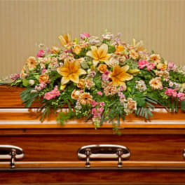 Wide casket spray of yellow lilies, peach and pink flowers, and green foliage on a wood casket
