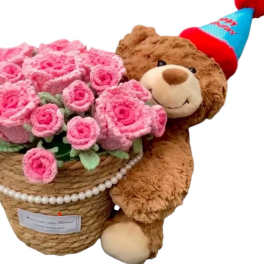 Pink rose bouquet in a woven basket beside a teddy bear wearing a party hat