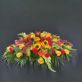 Elongated arrangement of yellow roses, orange gerberas, sunflowers, and red carnations with burgundy ribbons