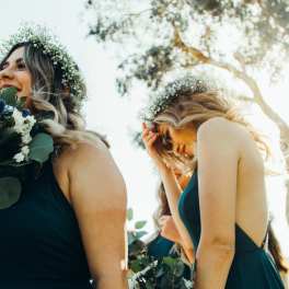 Bridesmaids in dark dresses holding bouquets and floral crowns outdoors