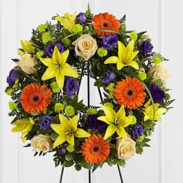 Circular floral wreath with yellow lilies, orange gerberas, cream roses, and purple blooms