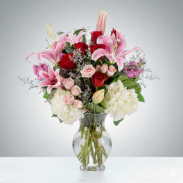 Tall mixed arrangement of pink lilies, red and blush roses, and white hydrangeas in a clear glass vase.