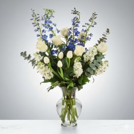 Tall arrangement of white roses, tulips, and blue delphinium in a clear glass vase