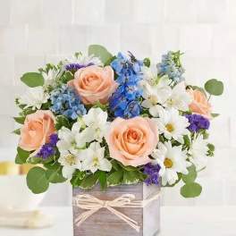 Peach roses and white daisies in a wooden box with blue flowers