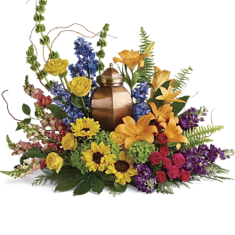 Colorful floral arrangement around a copper urn with lilies, roses, sunflowers, and blue blooms