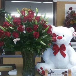 Red roses and lilies in a glass vase beside a white teddy bear with a red bow
