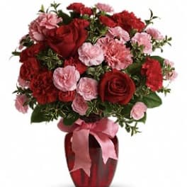 Red and pink roses and carnations arranged in a red glass vase with a pink ribbon bow