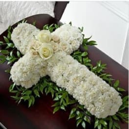 White floral cross with roses and carnations resting on greenery atop a dark wood casket