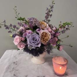 Lavender and pink rose arrangement in a white vase with a lit candle beside it.