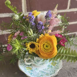 Mixed bouquet in a floral teacup and saucer with yellow, purple, pink, and white blooms