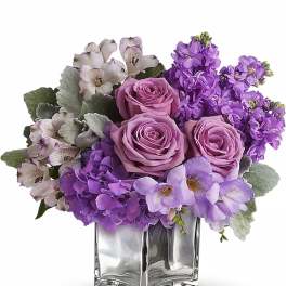 Purple roses and mixed lavender flowers in a clear square vase