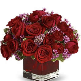 Red roses arranged in a square glass vase with small purple filler flowers