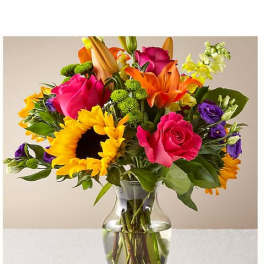 Colorful mixed bouquet in a clear glass vase with sunflowers, roses, lilies, and purple blooms