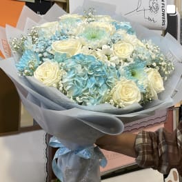 Large hand-tied bouquet of cream roses, blue hydrangeas, and blue and white mums wrapped in pale blue paper.