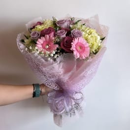 Handheld bouquet of pink gerberas, roses, and pale hydrangeas wrapped in lavender paper