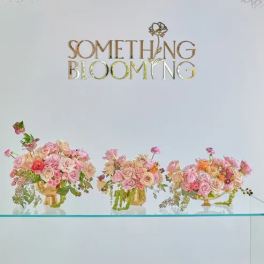 Three pink floral arrangements in gold vases beneath a "Something Blooming" sign