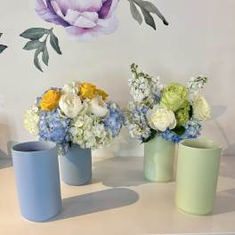 Two pastel floral arrangements in cylindrical vases with blue, white, and yellow blooms.
